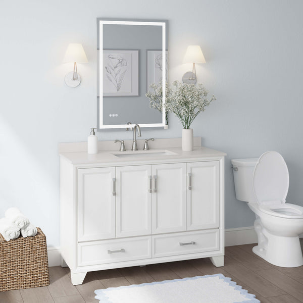 A modern bathroom with a white vanity, sink, and lighted mirror. Two wall sconces are above the vanity, with framed art and a vase of flowers. A toilet sits to the right, and a basket of towels is on the floor to the left.