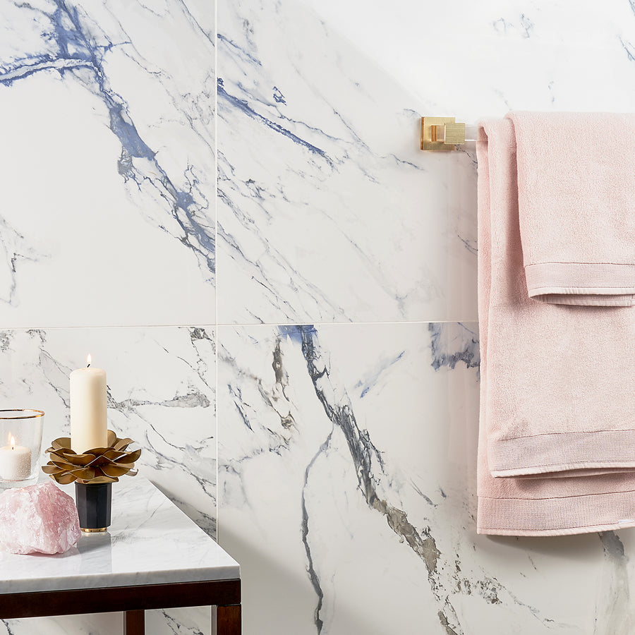 Bathroom scene with white marble walls, a gold towel bar holding folded pink towels, and a marble countertop displaying a lit candle, a pink crystal, and a gold decorative item.