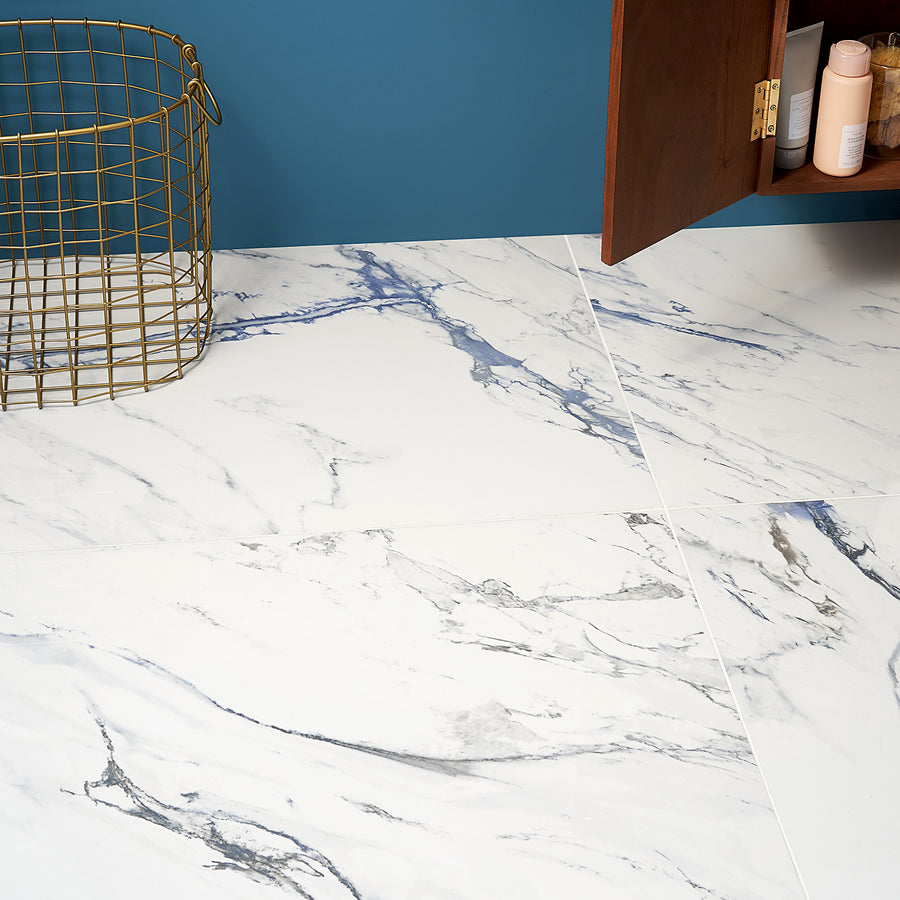White marble floor tiles with blue and gray veining next to a gold wire basket and a wooden cabinet against a teal wall. Some toiletries are visible inside the cabinet.
