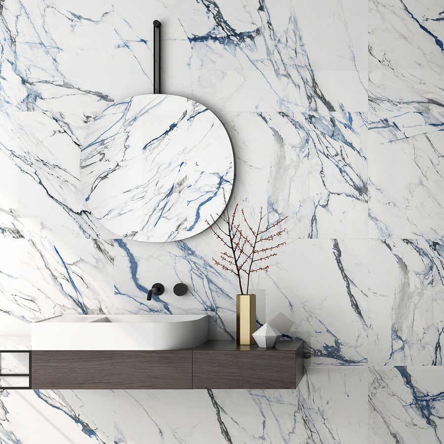 Modern bathroom with white and blue marble walls, a round mirror, minimalist floating sink with matte black fixtures, and a decorative branch in a vase on the countertop.