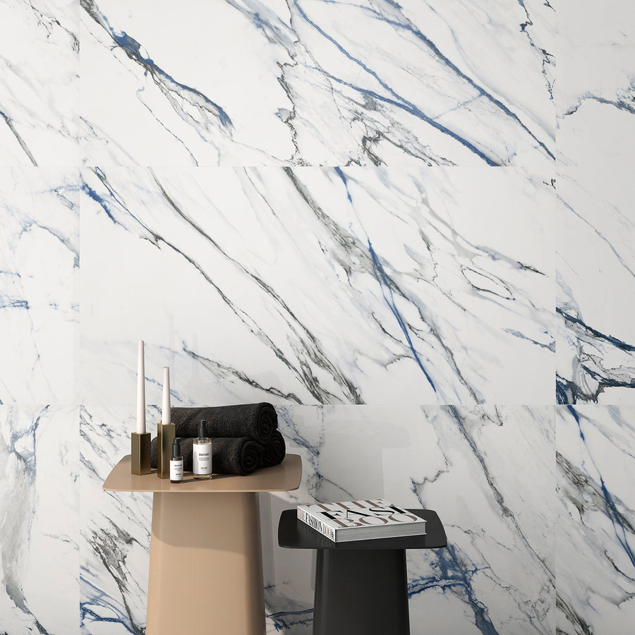 Two modern side tables in front of a white marble wall with gray and blue veining. One table holds candles, black towels, and skincare items; the other has a magazine on top.