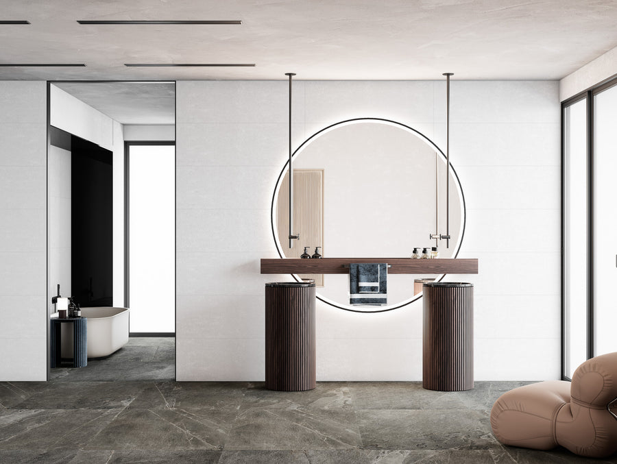 Modern bathroom with a large round backlit mirror, two pedestal sinks with dark wood finishes, minimalist fixtures, a taupe floor, and a glimpse of a bathtub in an adjacent room. Natural light enters from large windows.