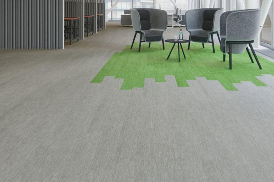 Modern office lounge with gray chairs around black tables on a green pixel-pattern carpet, surrounded by light gray Commercial Vinyl flooring. High tables and stools feature in the background.