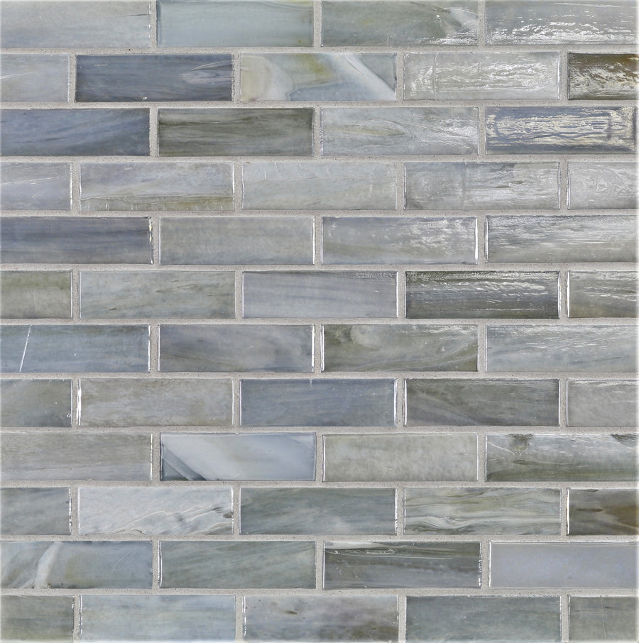 A close-up of a wall featuring Agate 1x3 Lucca Mosaic Glass Tile in staggered brick pattern, showcasing glossy, reflective rectangular tiles in varying shades of gray, blue, and green.