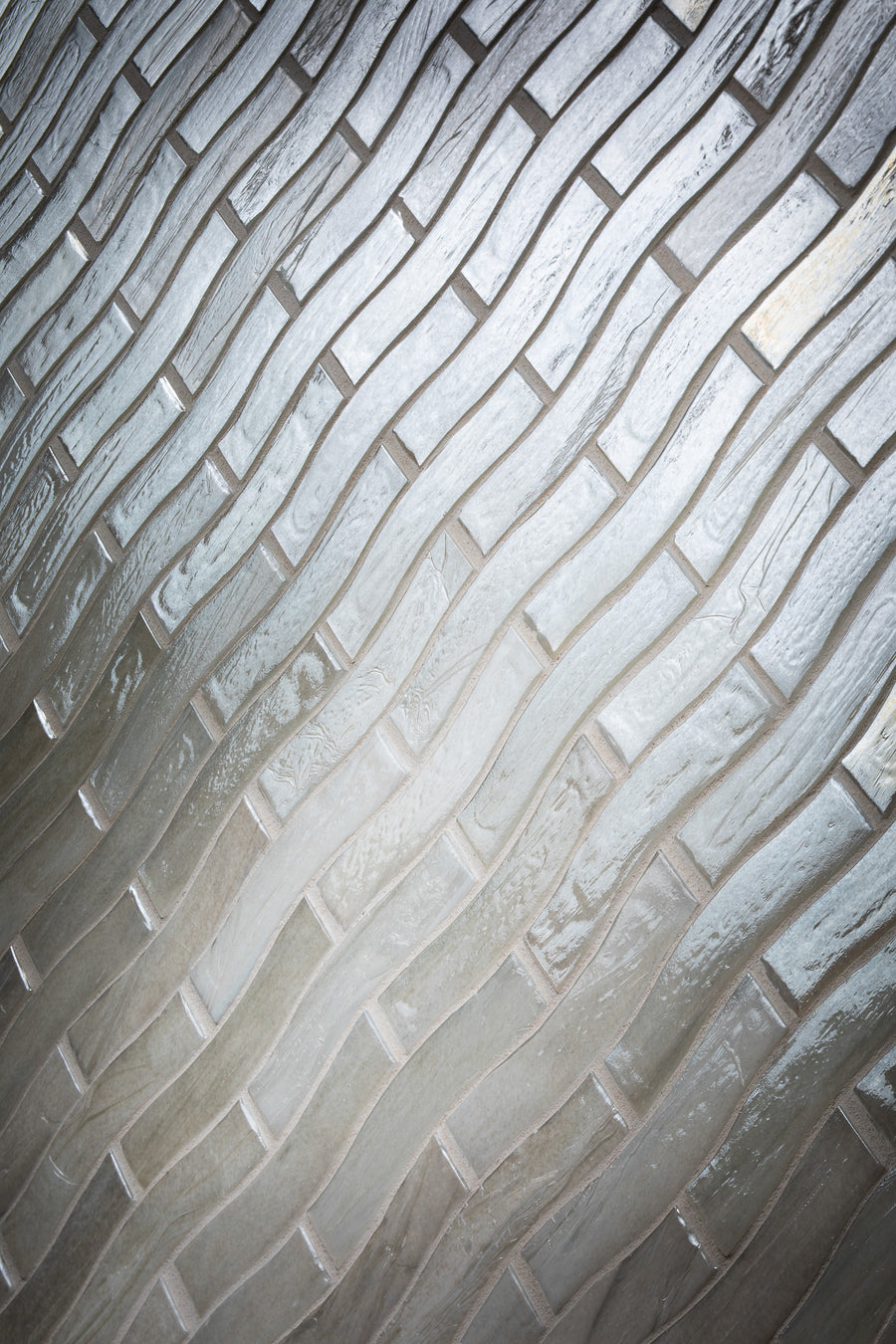 Wavy glass tiles arranged in a brick-like pattern with alternating light and dark shades, creating a textured and reflective surface.
