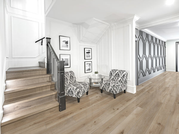 Modern hallway with light wood floors, a black and white patterned accent wall, two patterned armchairs, a round glass table, framed art on white walls, and a staircase with dark railing. Bright and minimalist decor.