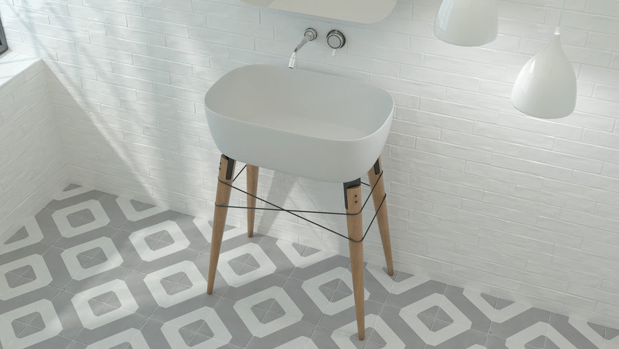 A modern white sink with wooden legs stands in a minimalist bathroom with white tiled walls and gray geometric patterned floor tiles. Two white pendant lights hang above the sink.