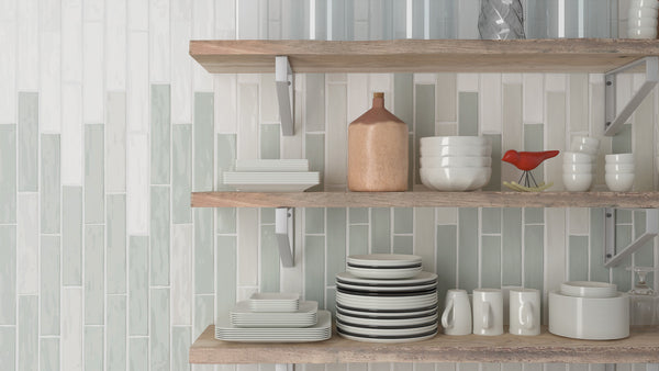 Three wooden kitchen shelves display neatly stacked white dishes, bowls, cups, a brown jug, a red bird figurine, and glassware, against a modern, vertically-tiled backsplash in light shades.