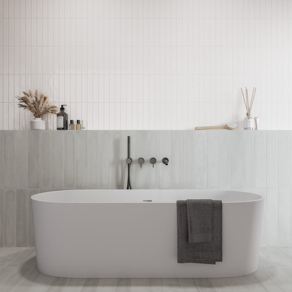 Minimalist bathroom with a modern white bathtub, a dark faucet, a gray towel draped over the tub, and decorative items like vases, bottles, and dried plants on a ledge against tiled walls.