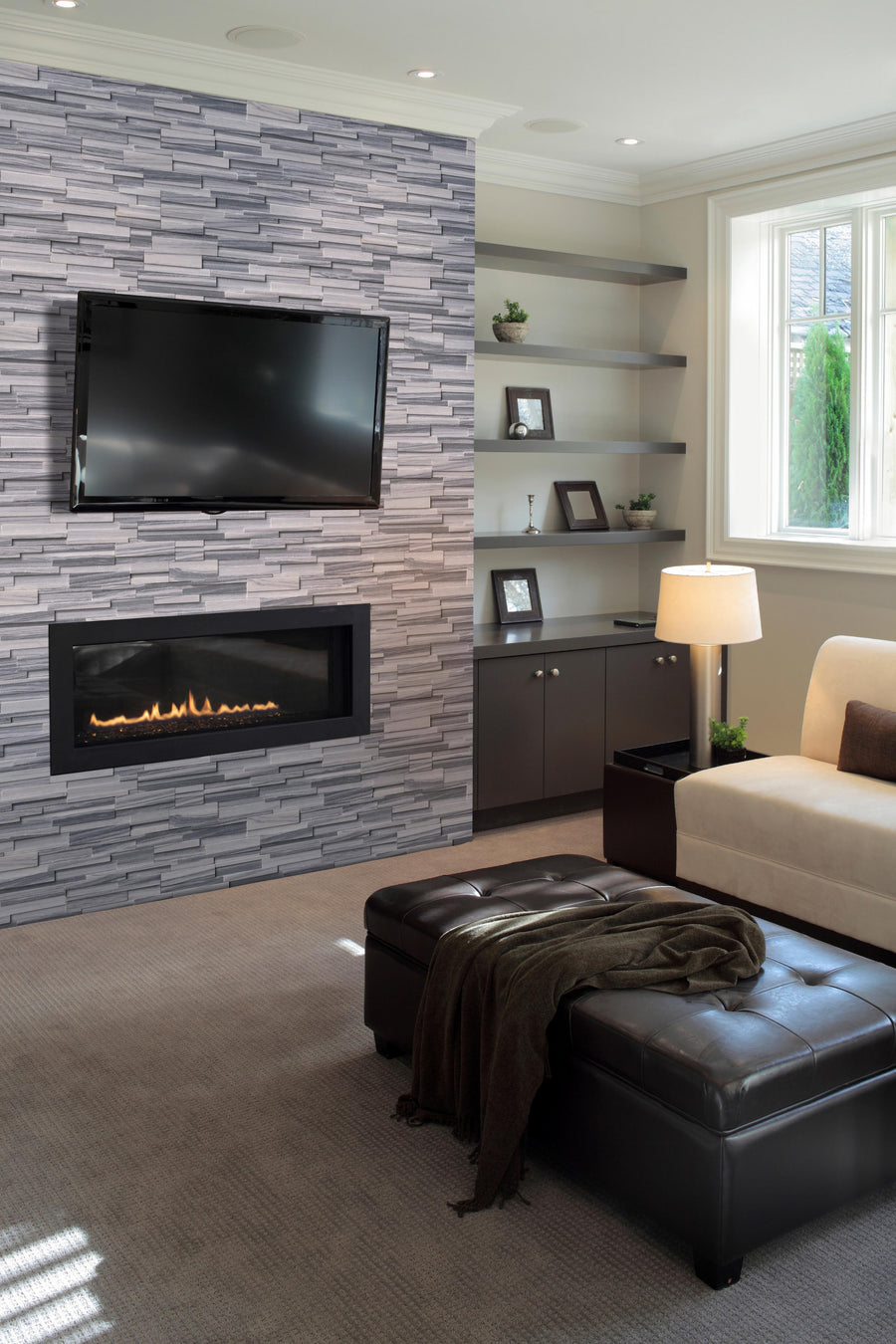 Modern living room with a wall-mounted TV above a sleek fireplace, gray stacked stone accent wall, built-in shelves with decorative items, a beige sofa, brown ottoman, and a large window letting in natural light.