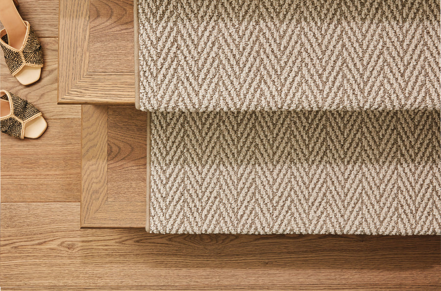 A pair of patterned sandals sits on a wooden floor next to carpeted stairs with a beige and white chevron pattern runner. The scene is viewed from above.