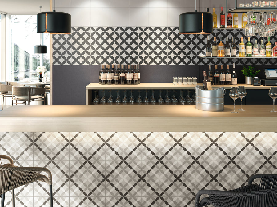 A modern bar with geometric patterned tiles on the counter and wall, black pendant lights, shelves with bottles and glasses, wine bottles displayed, and seating visible both inside and outside.