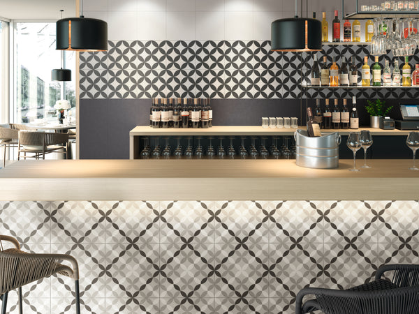 A modern bar with geometric patterned tiles on the counter and wall, black pendant lights, shelves with bottles and glasses, wine bottles displayed, and seating visible both inside and outside.