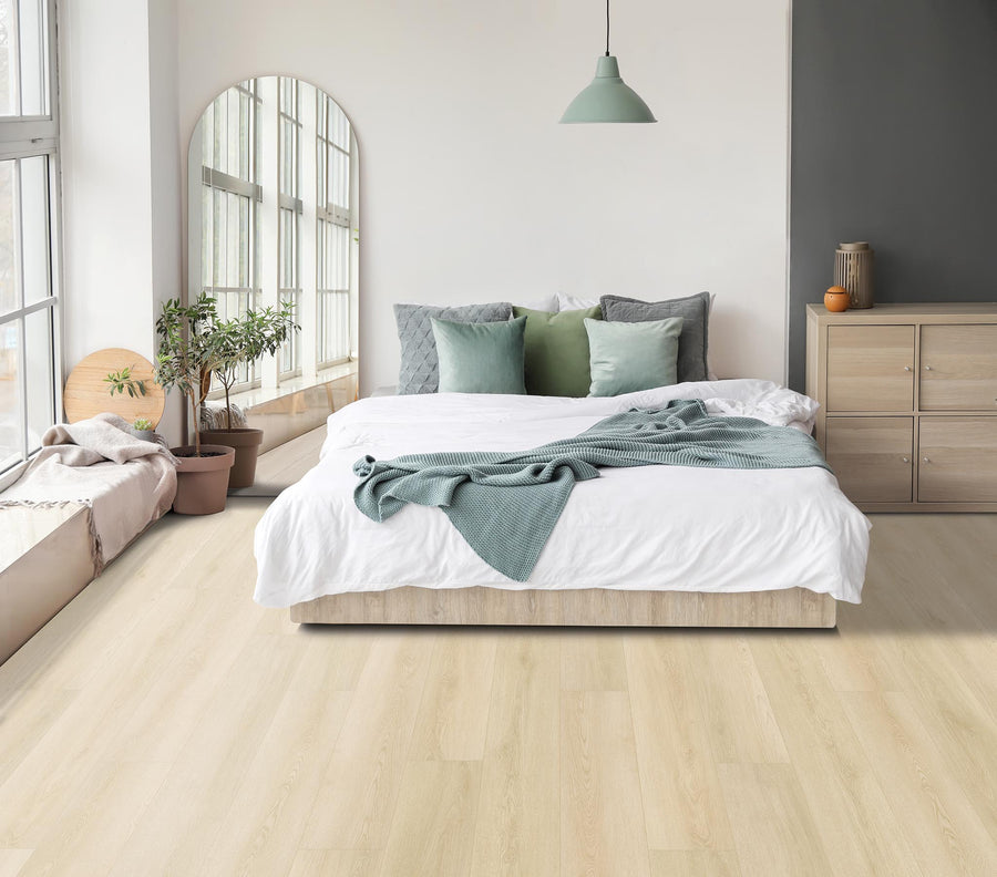 A modern bedroom with a large bed, green and gray pillows, a green blanket, wooden floors, potted plants, a floor mirror, and a dresser. Natural light streams in from tall windows on the left.