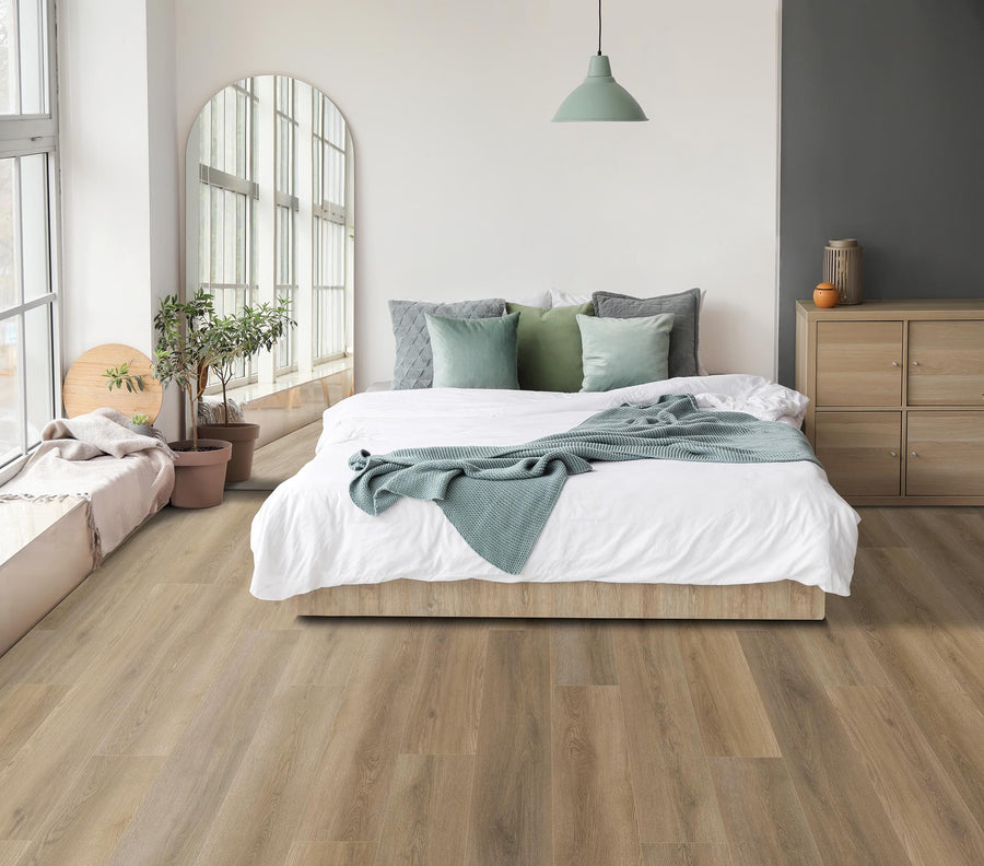 A modern bedroom with wood flooring, a large bed with white and green bedding, a light wood dresser, potted plants, a round mirror, and a pendant lamp, with sunlight streaming through tall windows.