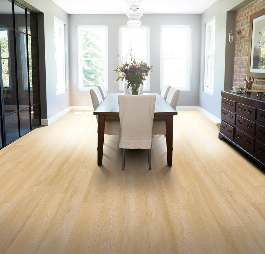Bright dining room with large windows, light wood flooring, a wooden table with six white upholstered chairs, a floral centerpiece, and a dark wooden sideboard against a brick accent wall.