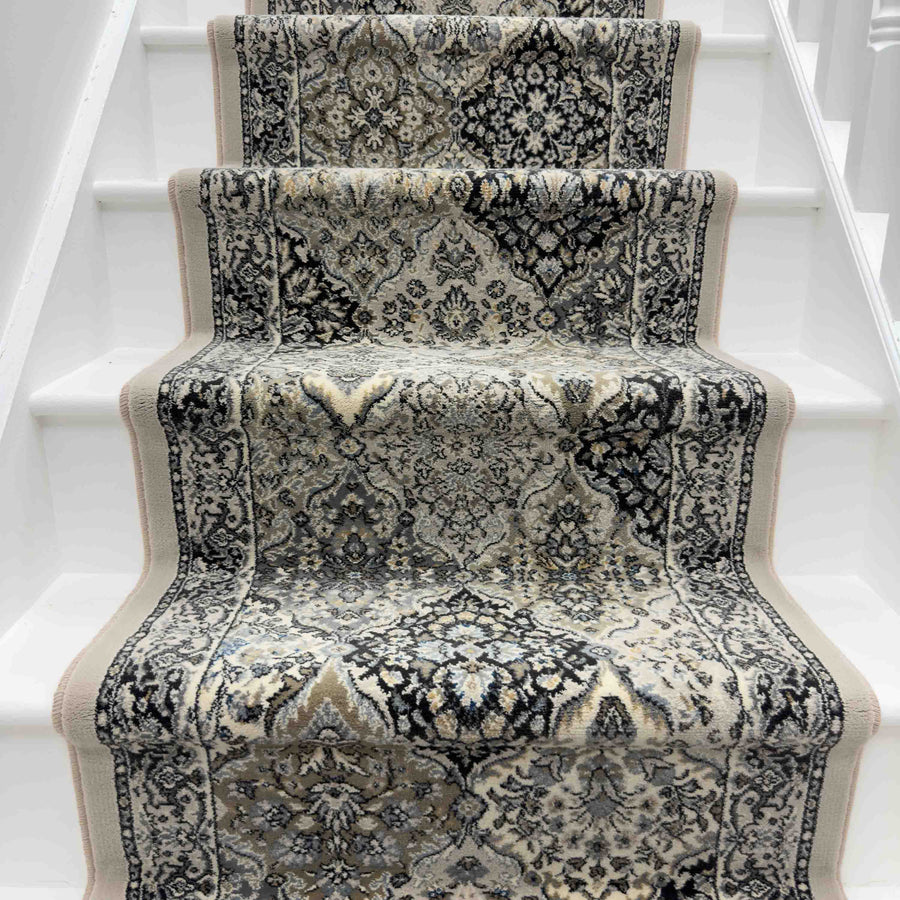 The Ancient Garden 57008-9696 Runner Rug, featuring intricate black, grey, and beige patterns, enhances the center of a white-painted staircase for a striking hallway runner look.