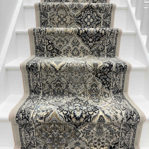The Ancient Garden 57008-9696 Runner Rug, featuring intricate black, grey, and beige patterns, enhances the center of a white-painted staircase for a striking hallway runner look.