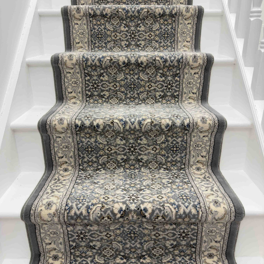A close-up of the Ancient Garden 57011-9666 Runner Rug, featuring gray, ivory, and blue designs on white wooden stairs, adds elegant style to any hallway or entryway.