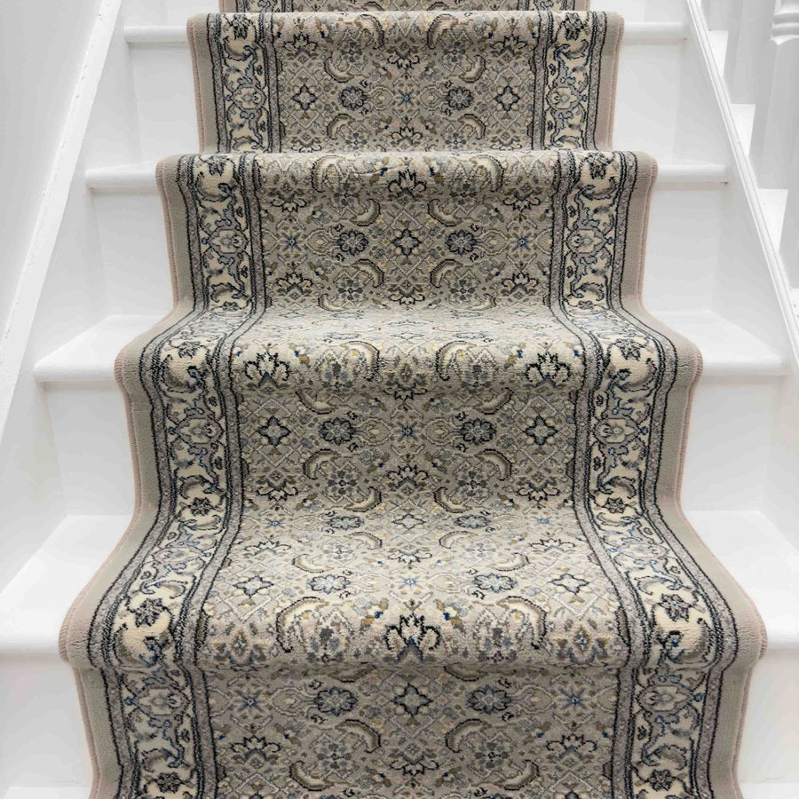 A staircase with white steps is styled with the Ancient Garden 57011-9666 Runner Rug, showcasing a beige, blue, and gray intricate traditional design that adds timeless elegance to your entryway.