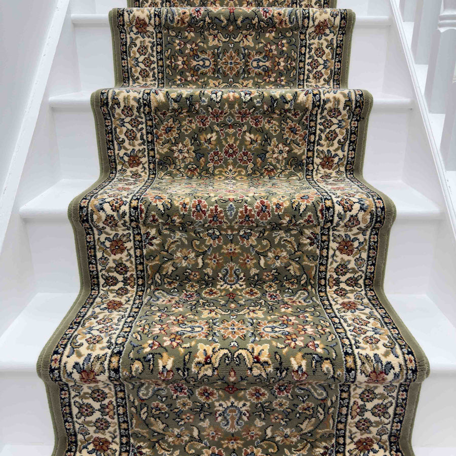 The Ancient Garden 57078-1414 Runner Rug, with floral and ornate designs in green, beige, and red, covers white wooden stairs. This hallway runner has a wavy edge that follows the contours of each step.