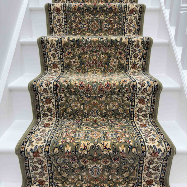 The Ancient Garden 57078-1414 Runner Rug, with floral and ornate designs in green, beige, and red, covers white wooden stairs. This hallway runner has a wavy edge that follows the contours of each step.