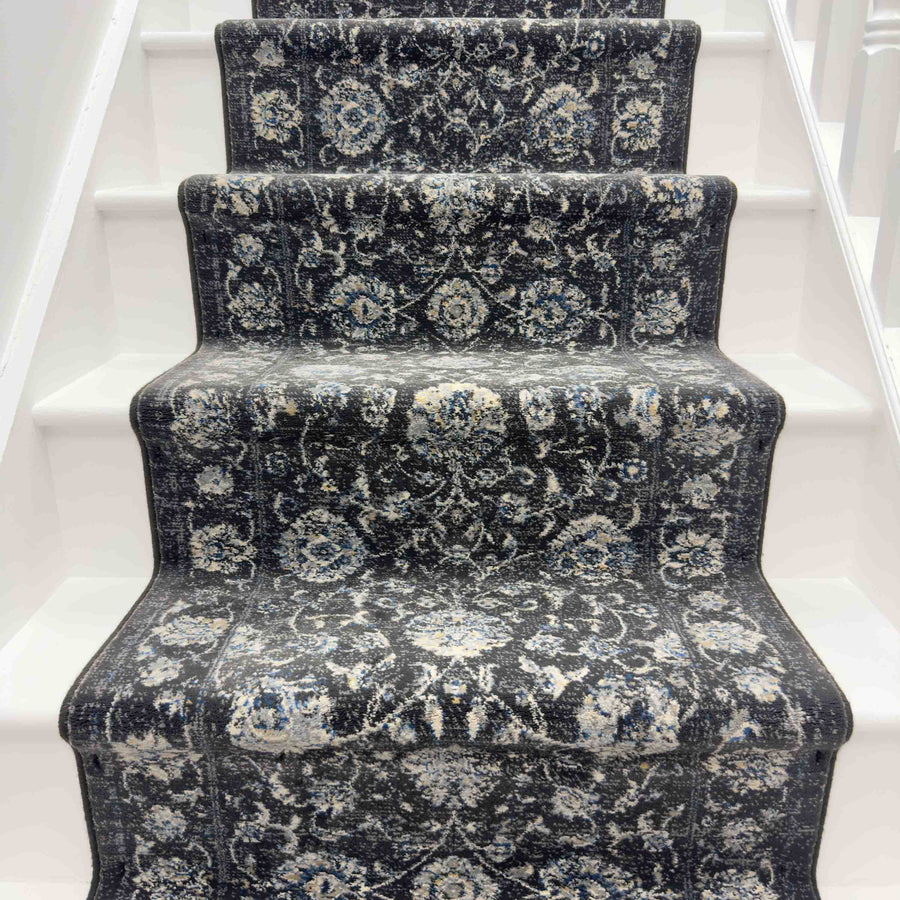 A staircase with white steps is covered by the Ancient Garden 3636 Runner Rug, a luxury rug displaying ornate floral and geometric patterns in shades of gray, blue, and cream.
