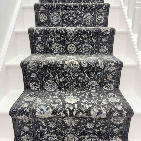 A staircase with white steps is covered by the Ancient Garden 3636 Runner Rug, a luxury rug displaying ornate floral and geometric patterns in shades of gray, blue, and cream.