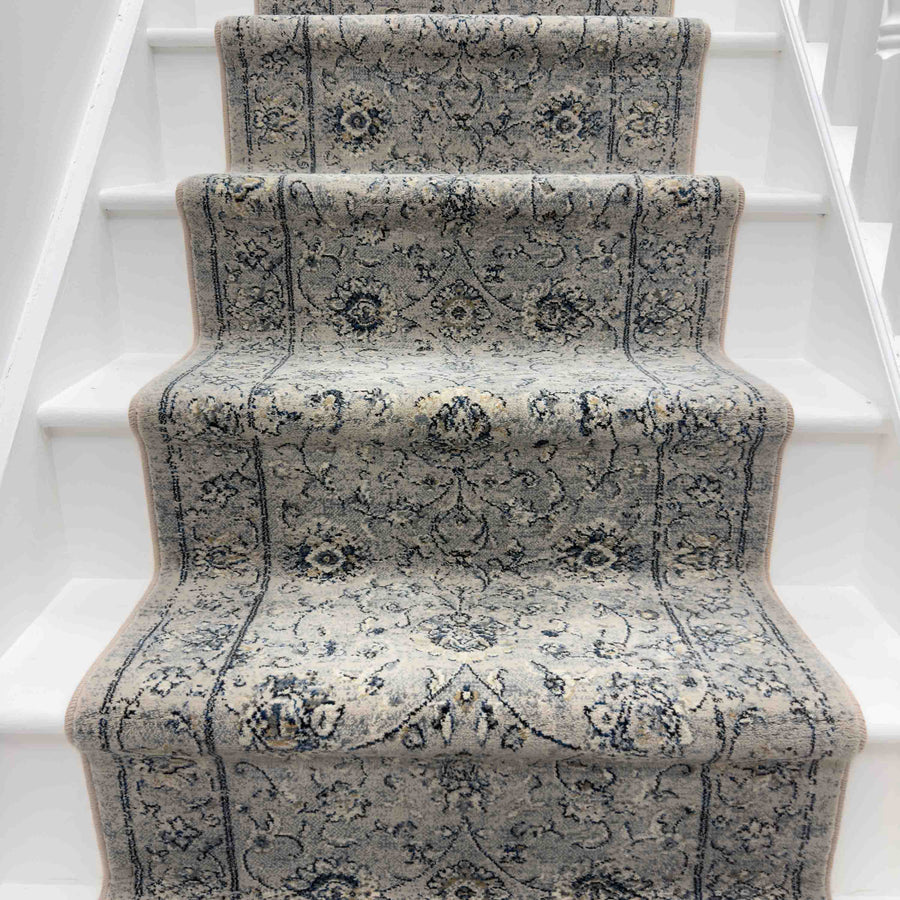 The Ancient Garden 57126-6666 Runner Rug, featuring a light gray and blue ornate design, covers the center of a white staircase with visible steps and side panels, making it an ideal hallway runner.