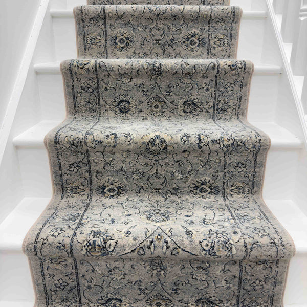 The Ancient Garden 57126-6666 Runner Rug, featuring a light gray and blue ornate design, covers the center of a white staircase with visible steps and side panels, making it an ideal hallway runner.