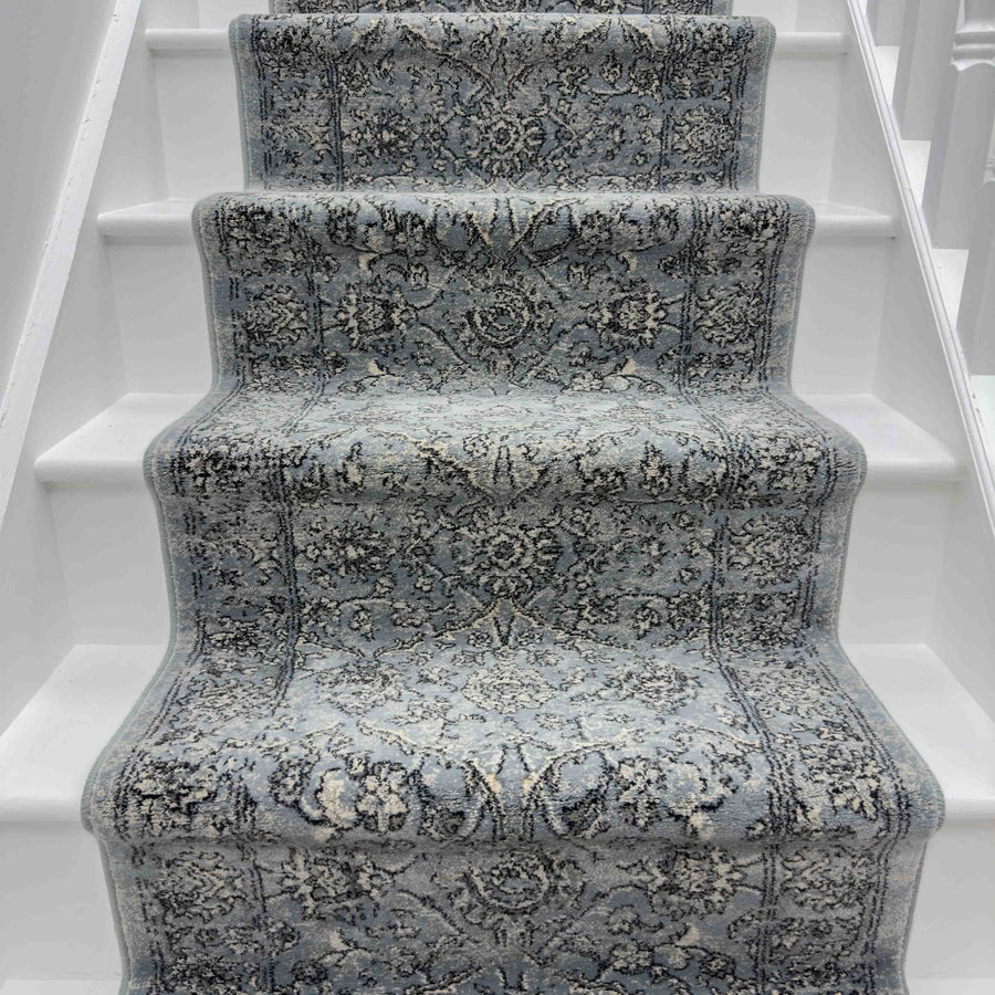 A staircase with white steps features the Ancient Garden 57136-4646 Runner Rug—a blue and gray hallway runner with intricate floral patterns.