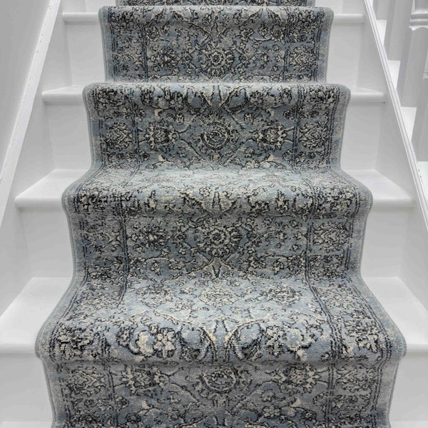 A staircase with white steps features the Ancient Garden 57136-4646 Runner Rug—a blue and gray hallway runner with intricate floral patterns.