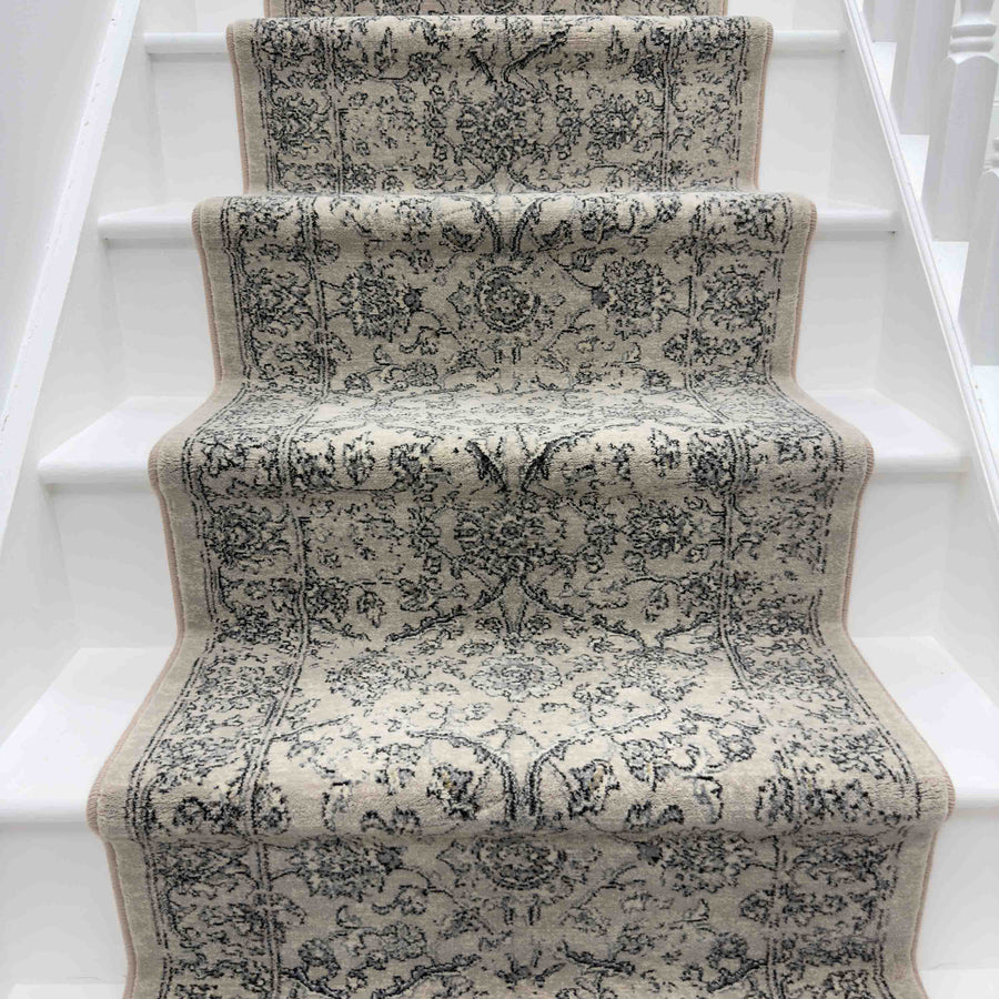 A close-up of a white staircase highlights the elegant Ancient Garden 57136-4646 Runner Rug, featuring intricate beige and gray floral and geometric designs covering the center of the steps.