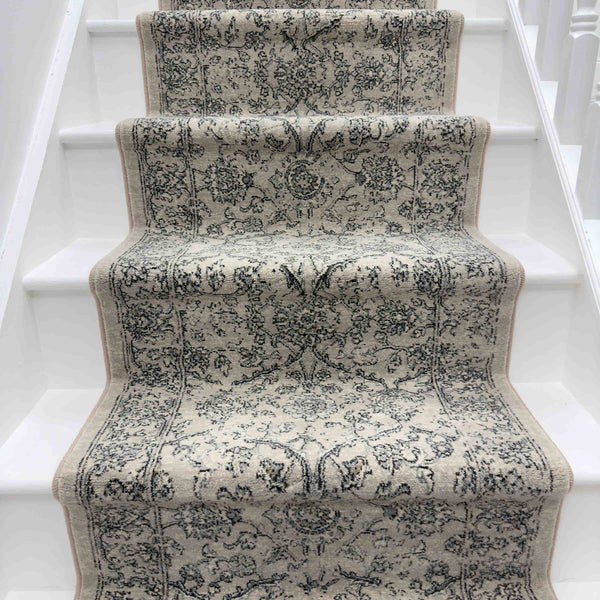 A close-up of a white staircase highlights the elegant Ancient Garden 57136-4646 Runner Rug, featuring intricate beige and gray floral and geometric designs covering the center of the steps.
