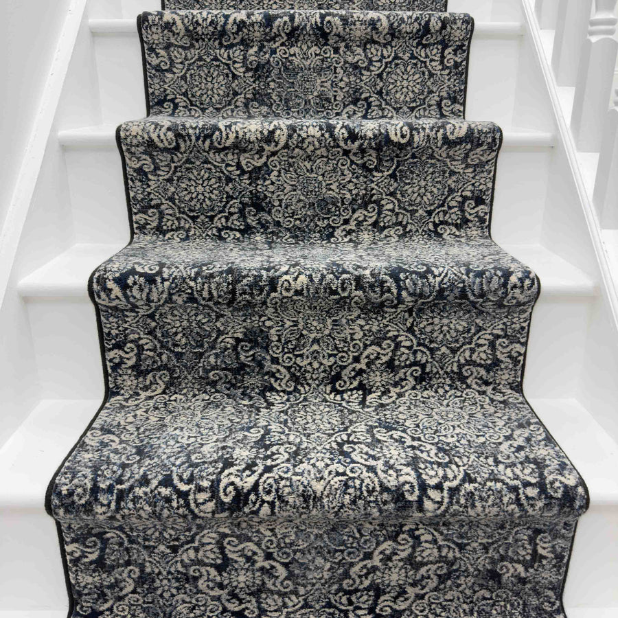 The Ancient Garden 57162-3696 Runner Rug, in black and white with an ornate design and elegant black border, covers the white wooden staircase steps.
