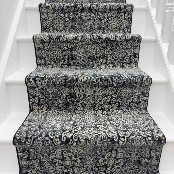 The Ancient Garden 57162-3696 Runner Rug, in black and white with an ornate design and elegant black border, covers the white wooden staircase steps.