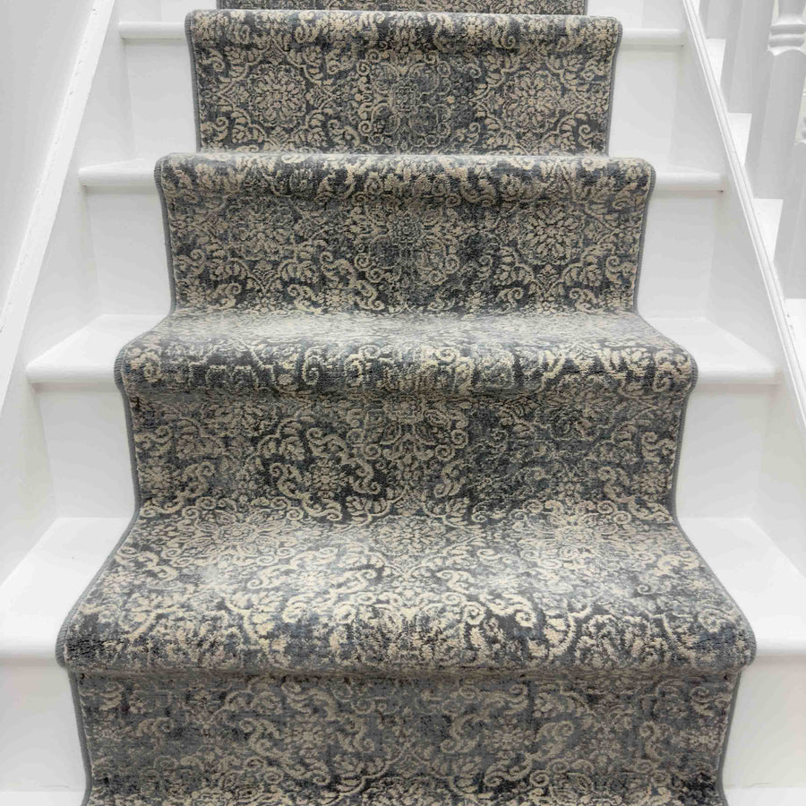 A staircase with white steps is adorned by the Ancient Garden 57162-3696 Runner Rug in gray and cream, showcasing intricate, ornate patterns for an elegant look.