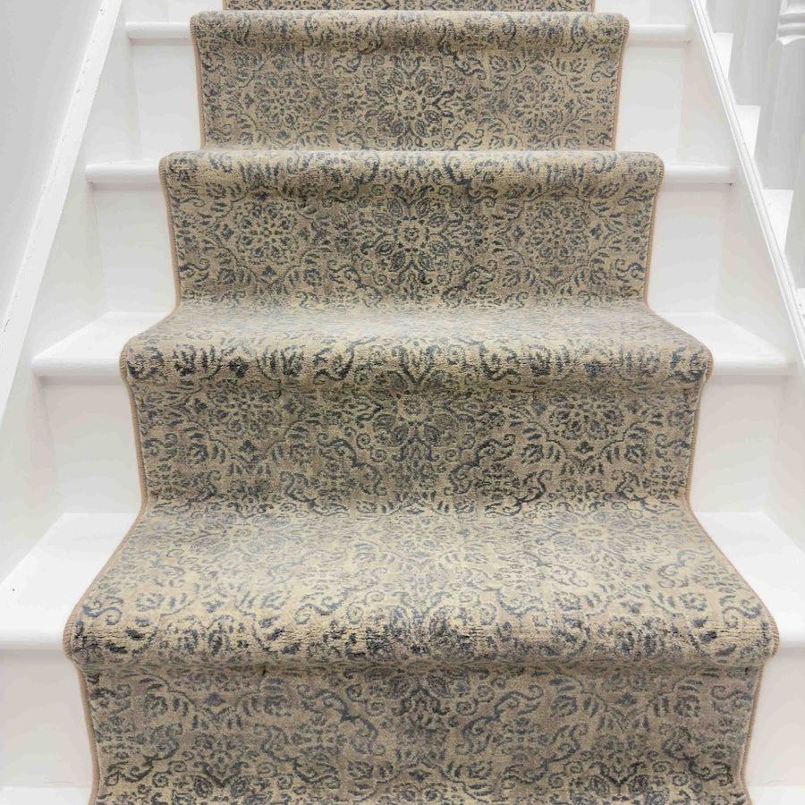 A staircase with white steps features the Ancient Garden 57162-3696 Runner Rug in beige and gray, showcasing an intricate, symmetrical pattern for a refined, classic look up the center of the hallway.