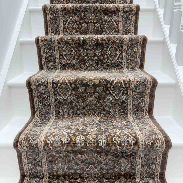 A brown, floral-patterned Ancient Garden 57276-9295 Runner Rug covers the white steps of a narrow staircase, curving over each edge. White railings line both sides.