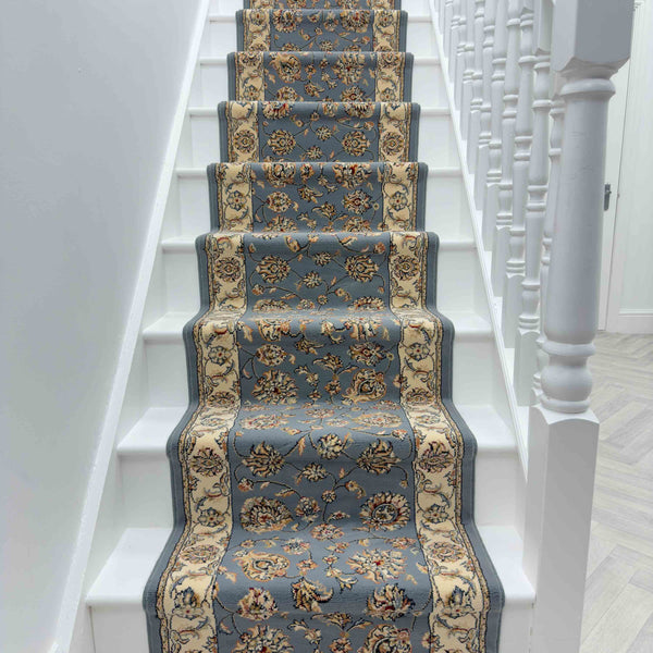 A staircase with white steps and railings features the Ancient Garden 57365-5464 Runner Rug, showcasing a blue background with beige floral patterns down the center for an elegant touch.