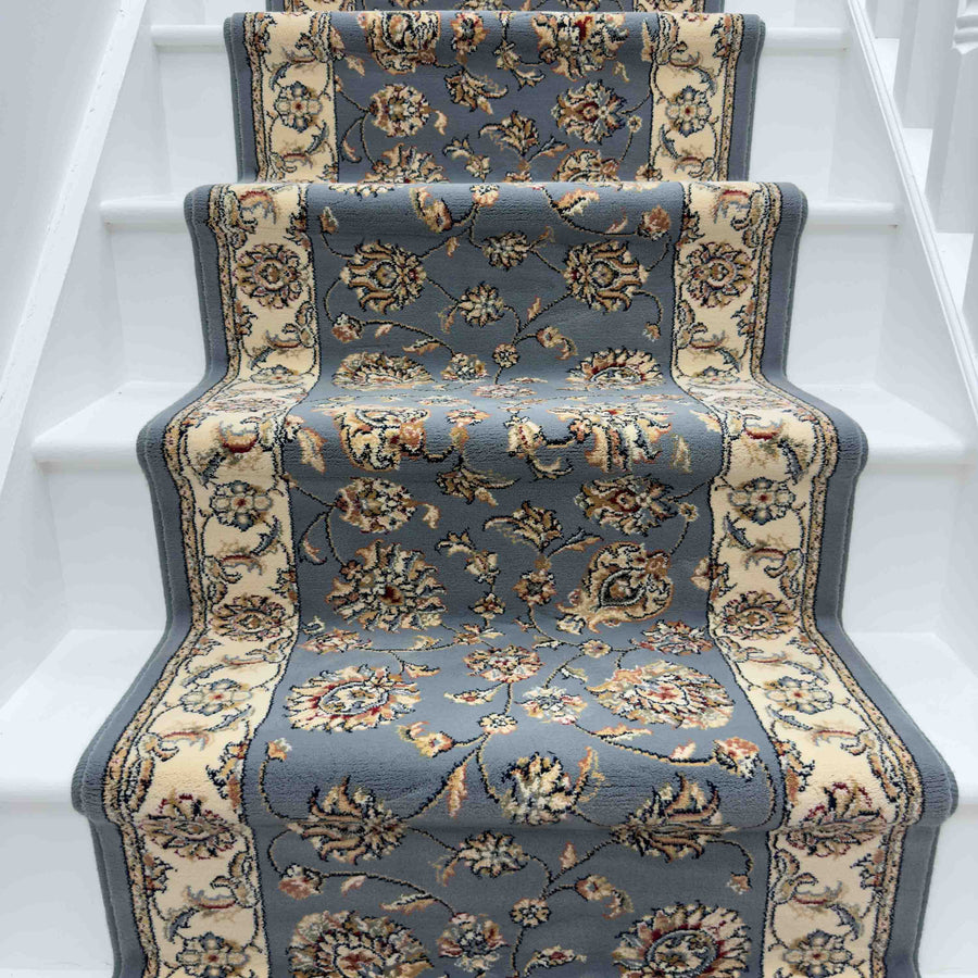 A close-up of a staircase showcases the Ancient Garden 57365-1464 Runner Rug, featuring elegant floral and ornate blue, beige, and cream designs across the white-painted steps.