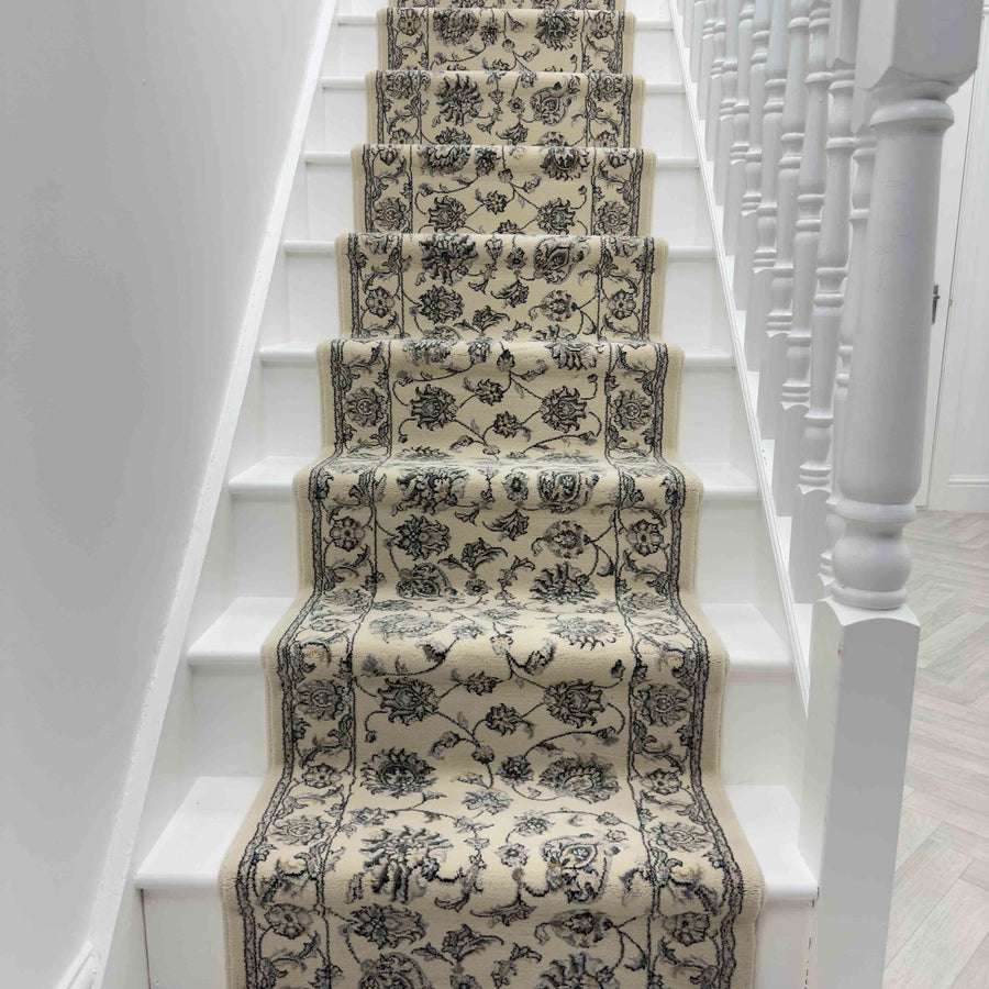 A staircase with white steps and spindles is accented by the Ancient Garden 57365-6666 Runner Rug, showcasing a luxurious beige design with a black floral pattern up the center.
