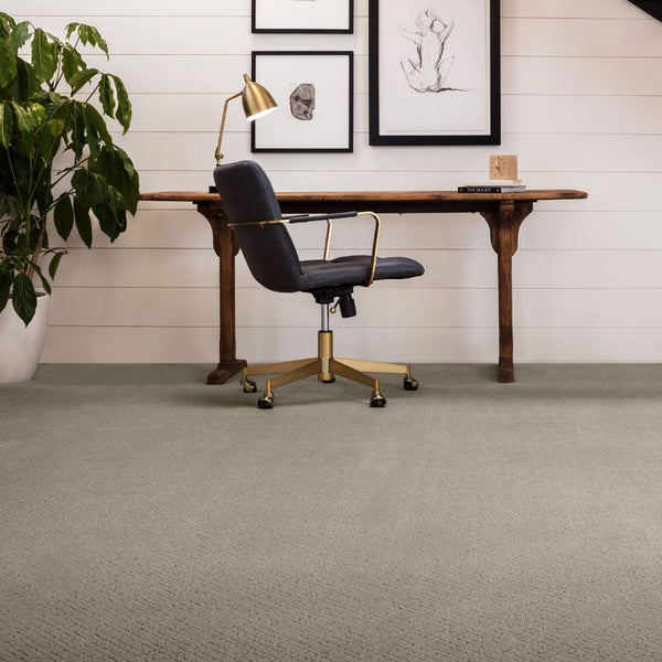 A wooden desk with a dark chair on wheels sits in a modern, minimal home office with light carpet, white shiplap walls, framed art, a potted plant, and a gold desk lamp.