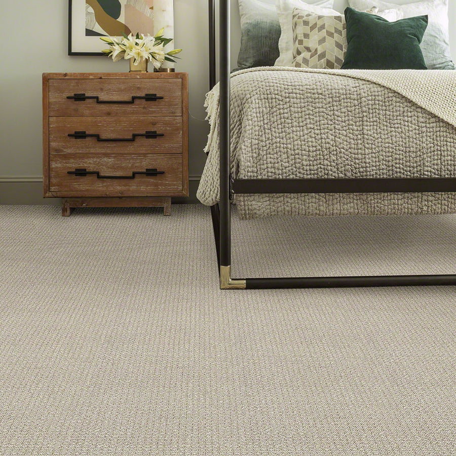 A cozy bedroom with a metal-framed bed, textured knit bedding, green and gray pillows, a wooden nightstand with three drawers, a vase of white flowers, and a soft, light-colored carpet.