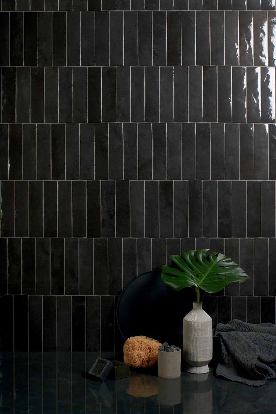 A glossy black tile wall with rectangular tiles arranged vertically. In front, there is a green monstera leaf in a white vase, a dark plate, a sponge, a small stone bowl, and a folded dark towel on a reflective surface.