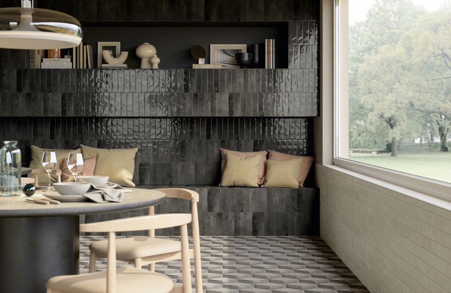 A modern dining area with light wooden chairs and a round table set for a meal. Behind, a built-in dark tiled bench with beige pillows sits beneath shelves holding books and decorative objects. Large window overlooks greenery.