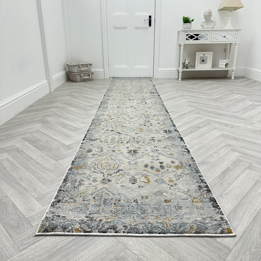 The Analise 7602-199 Runner Rug, featuring grey, beige, and gold patterns, lies on light herringbone wood in a hallway with a white door, decorative console table, and charming wicker basket.
