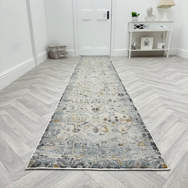 The Analise 7602-199 Runner Rug, featuring grey, beige, and gold patterns, lies on light herringbone wood in a hallway with a white door, decorative console table, and charming wicker basket.