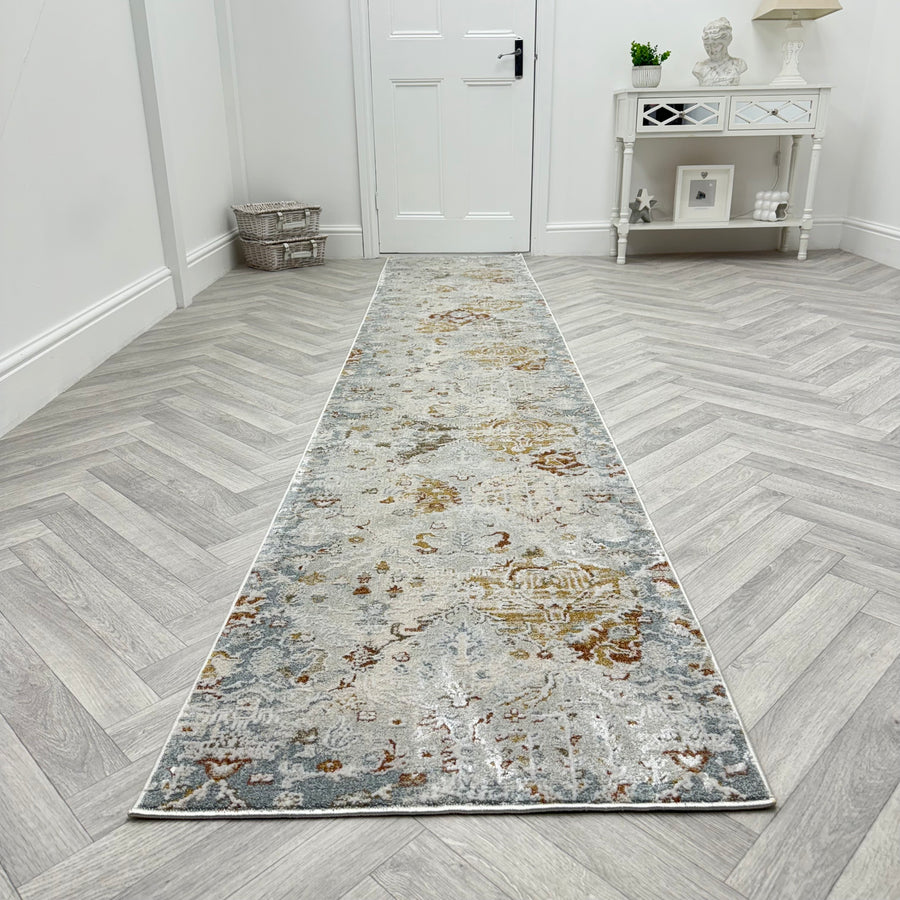 The Analise 7602-199 Runner Rug, featuring muted blue, beige, and brown patterns, lies on a light gray herringbone floor in a bright hallway with white walls, a door, console table, and baskets.