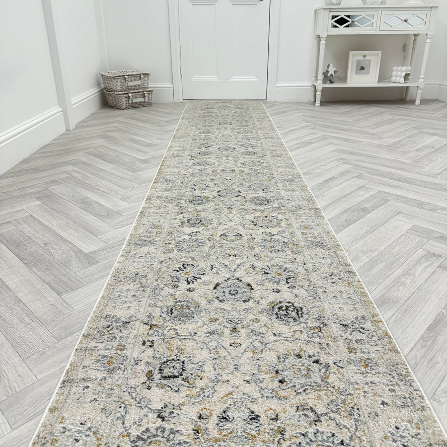 The Analise 7604-580 Runner Rug, featuring a long, patterned design in grey and beige, is laid on a light herringbone wood floor in a bright hallway with a white door, basket, and mirrored console table in the background.